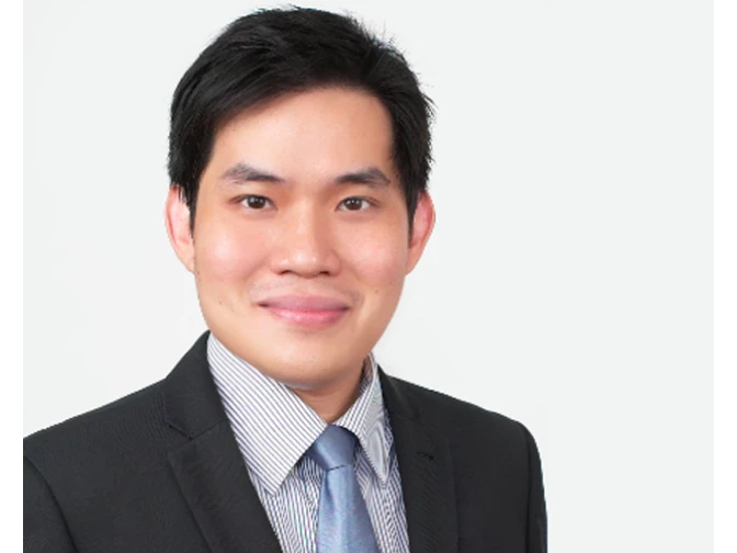 A man with short black hair and a pleasant smile, wearing a black suit jacket, a fine-checkered shirt, and a blue tie. He is posed against a solid, bright white background.