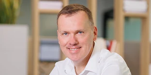 A man with short brown hair and blue eyes, smiling at the camera while wearing a white button-down shirt. The background is a bright, blurred office with wooden shelving and a hint of greenery.