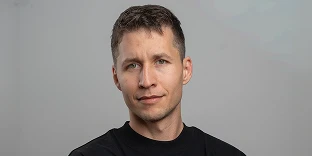 A man with short, dark hair and green eyes, looking directly at the camera with a neutral, professional expression, wearing a black crew-neck shirt against a solid grey background.
