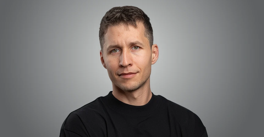 A man with short, dark hair and green eyes, looking directly at the camera with a neutral, professional expression, wearing a black crew-neck shirt against a solid grey background.